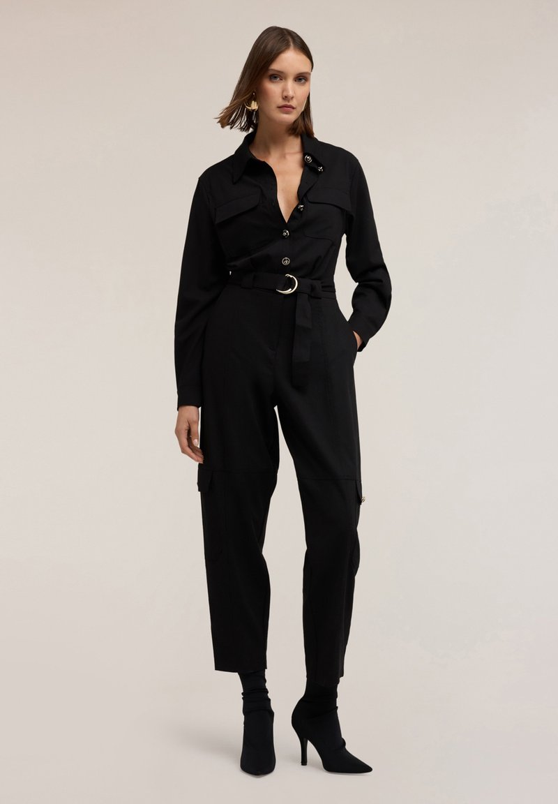 Zwarte jumpsuit met lange mouwen, knoopsluiting aan de voorkant en een tailleband. Bevat twee borstzakken en zijzakken in cargostijl. Gladde textuur.