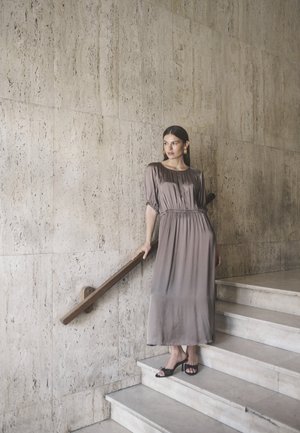 Maxikleid aus weichem taupefarbenem Stoff mit kurzen Puffärmeln, gerafftem Taillenbereich und fließender Silhouette, kombiniert mit schwarzen offenen High Heels.