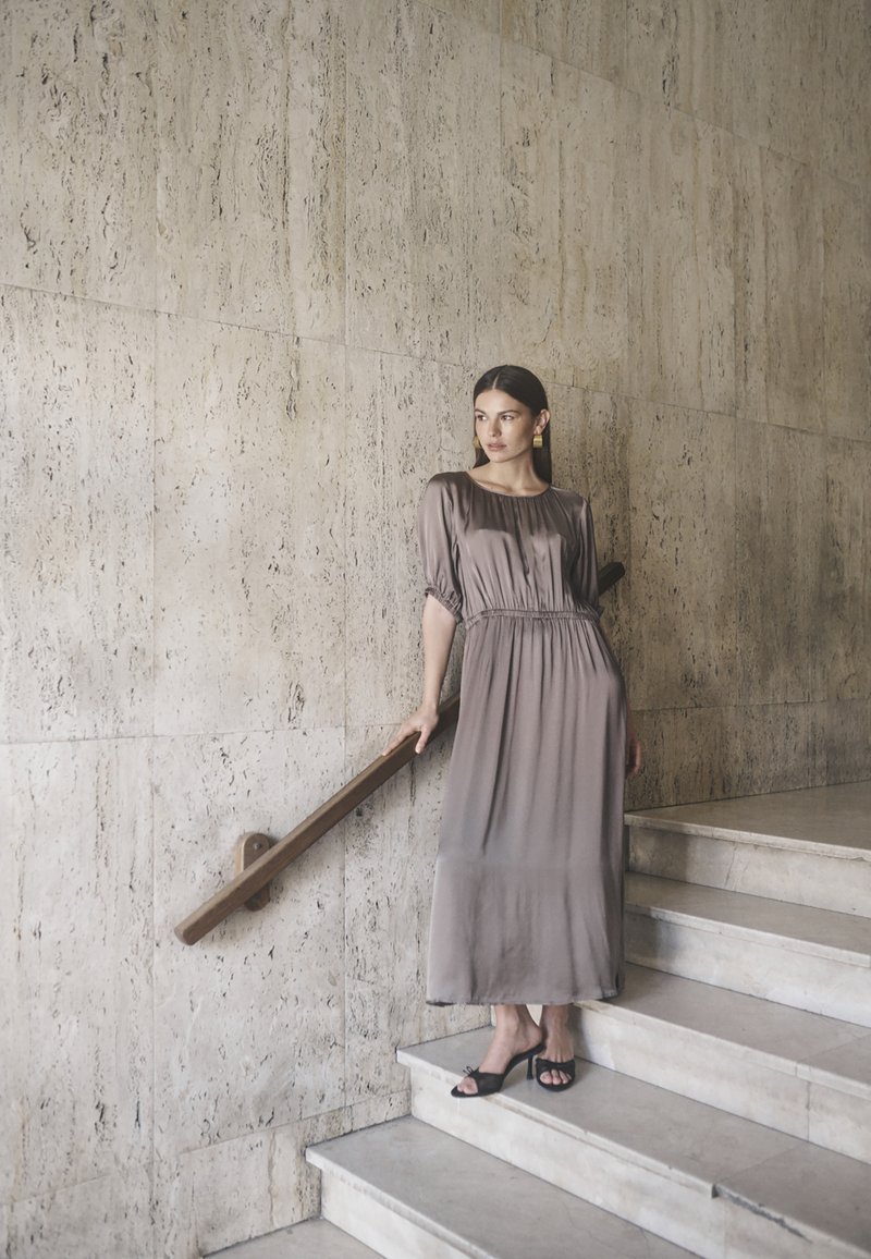 Maxikleid aus weichem taupefarbenem Stoff mit kurzen Puffärmeln, gerafftem Taillenbereich und fließender Silhouette, kombiniert mit schwarzen offenen High Heels.