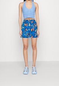 Světle modrý pletený halter top na cvičení v kombinaci s modrými květinovými šortkami, které mají jasně barevné květy. Modré sandály na podpatku celý vzhled doplňují.