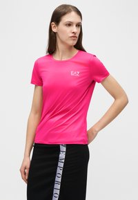 Γυναίκα που φοράει ένα φωτεινό ροζ κοντομάνικο πουκάμισο EA7 Emporio Armani και μαύρη φούστα με κάθετη λωρίδα με το λογότυπο Emporio, κοιτάζοντας προς τα αριστερά.