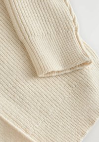 Pull en tricot crème avec des côtes verticales et une finition texturée. Le tissu semble doux et légèrement duveteux, avec une coupe décontractée.