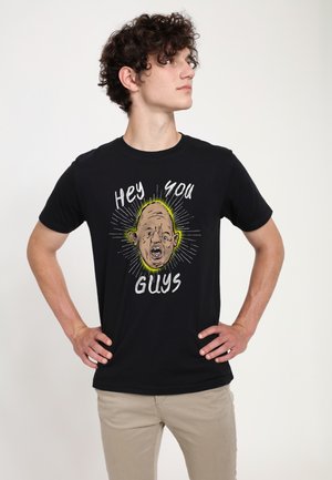 Ung mand med krøllet hår står med hænderne på hofterne iført en sort T-shirt med en tegneserieansigt og teksten "Hey You Guys."