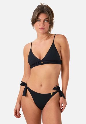 Kobieta o jasnej cerze i związanych z tyłu włosach, ubrana w czarne bikini z wiązaniami po bokach i małym złotym emblematem na gładkim białym tle.