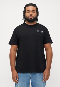 Man met gevlochten haar en baard, gekleed in een zwart T-shirt met witte tekst "J&J since 1990 Society club" en blauwe jeans, staande tegen een witte achtergrond.