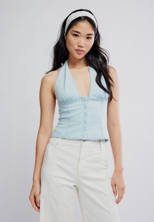 Mladá žena s dlhými tmavými vlasmi, ktorá nosí svetlomodrý zapínaný halter top a biele nohavice s vysokým pásom na jednofarebnom pozadí.