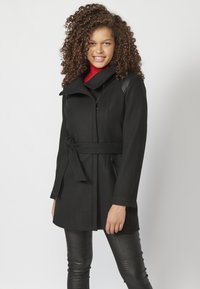 Cappotto nero con vita cinturata e ampio colletto, dotato di tasche con zip e dettagli sulle spalle. Modella che indossa pantaloni neri aderenti e strutturati.