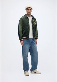 Πράσινο σατέν μπουφάν τύπου μπόμπερ με κεντημένο μετάλλιο, μανσέτες με ρίγες, λευκό μπλουζάκι, ανοιχτό μπλε τζιν σε φαρδιά γραμμή και κρεμ αθλητικά παπούτσια.