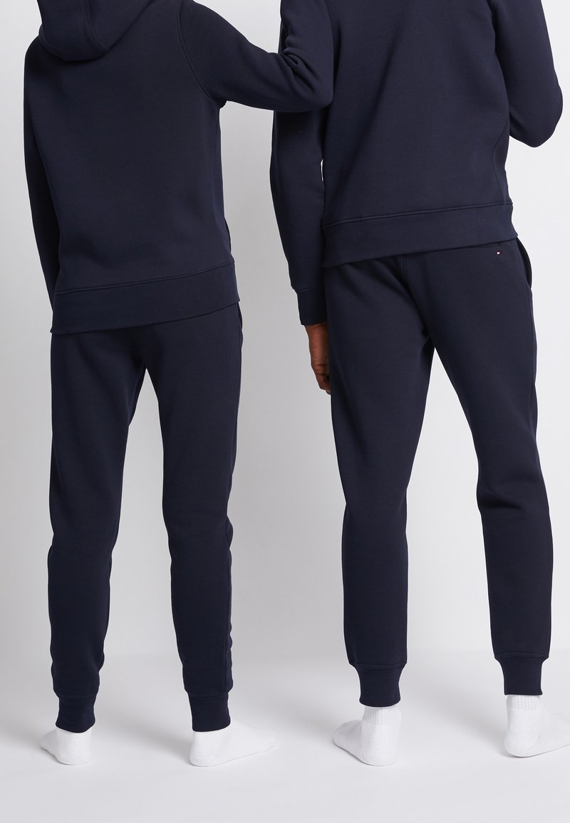 Deux personnes sont debout côte à côte, portant des sweats à capuche bleu marine assortis et des pantalons de jogging avec des chaussettes blanches, vues de dos.