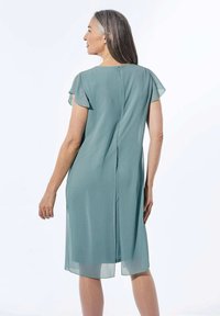 Een teal jurk tot de knie, gemaakt van lichte stof, met korte vlindermouwen en een ritssluiting op de rug. De jurk heeft een verticale slit voor bewegingsvrijheid.