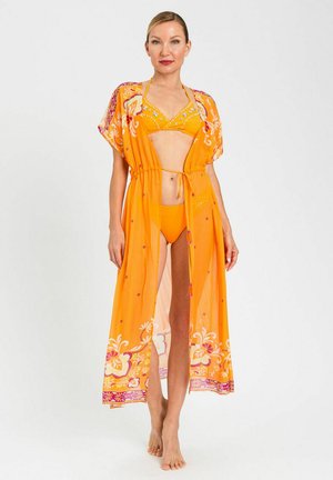 Donna che indossa un bikini arancione brillante con un copricostume floreale trasparente abbinato, in piedi scalza su uno sfondo bianco uniforme.