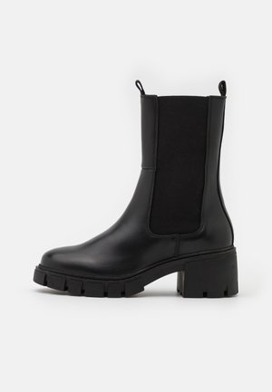 Botte noire en cuir jusqu'au milieu du mollet avec semelle crantée épaisse, talon bloc, panneaux latéraux élastiques et languettes de tirage sur un fond clair uni.