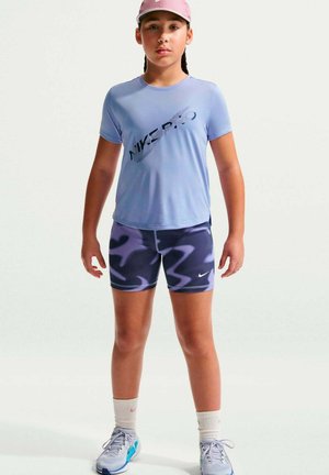 Junge Person trägt ein lila Nike Pro T-Shirt, gemusterte Sportshorts, weiße Socken, graue Sneakers und eine pinke Kappe und steht nach vorne gerichtet.