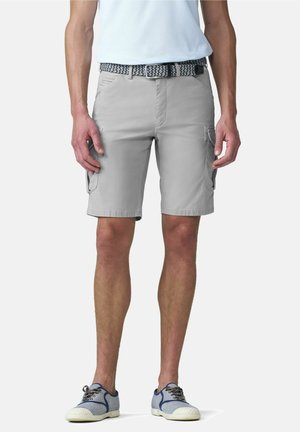 Man draagt lichtgrijze cargoshorts, een patroonriem, een wit shirt en blauwe casual sneakers, staand tegen een effen achtergrond.