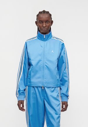 Person mit geflochtenem Haar, die einen blauen glänzenden Trainingsanzug mit weißen und schwarzen Streifen und einem kleinen weißen Air-Jordan-Logo auf der Jacke trägt.