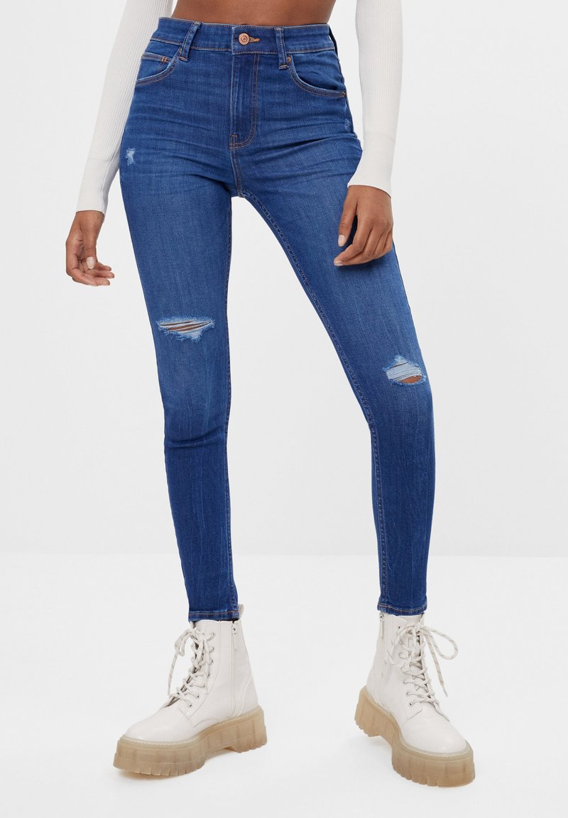 Jeans skinny de mezclilla azul de cintura alta con detalles desgastados en las rodillas. Combinados con botas de combate blancas con cordones y suela gruesa.
