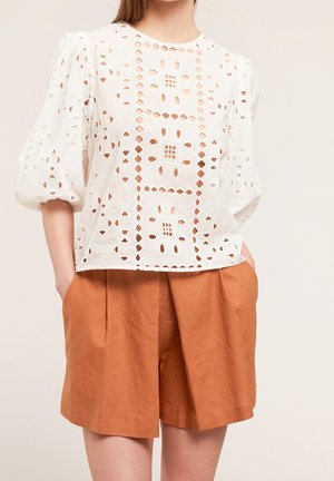 Kvinde iført hvid øjlet blonderbluse med puffede ærmer og højtaljede rustfarvede plisserede shorts med hænderne i lommerne.