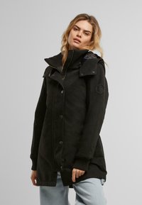 Manteau noir long avec un col haut, des boutons de fermeture et des poignets côtelés. Comprend une capuche et un marquage subtil sur la manche.