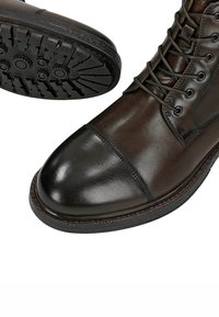Stivali in pelle marrone scuro con punta a cappuccio liscia, suola in gomma nera testurizzata e design tradizionale con lacci. Dettagli di cucitura rinforzata visibili.