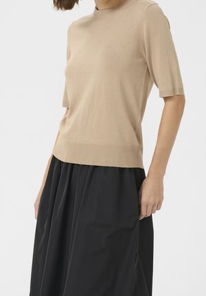 Woman wearing a beige short-sleeve knit top and a high-waisted black skirt standing against a plain background.