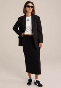 Fekete szabott zakó egy fehér crop top felett, fekete logóval, párosítva egy fekete midi szoknyával és fekete sportcipővel, fehér csíkokkal.