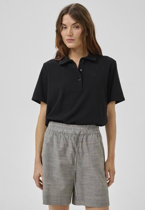 Nő fekete rövid ujjú polóinget és magasderekú, szürke-fehér csíkos rövidnadrágot visel, szürke háttér előtt.