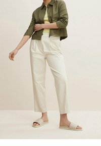 Giacca cropped verde oliva, top giallo a fantasia, pantaloni bianchi a gamba larga e sandali leggeri beige trapuntati su uno sfondo neutro.