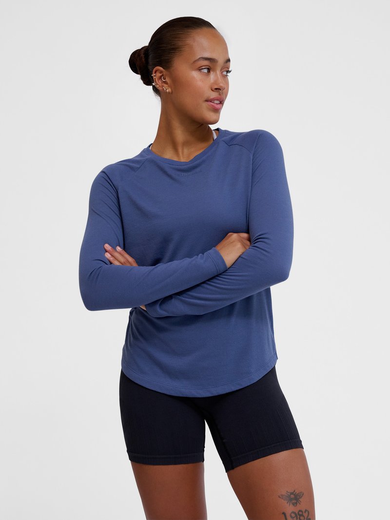 Frau mit Dutt, trägt blaues Langarmshirt und schwarze Shorts, Arme verschränkt, schaut nach links vor einfachem Hintergrund.