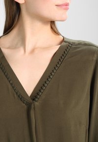 Olivgrön blus med V-ringning och texturerad pom-pom kant, tillverkad av mjukt material med en slät yta.