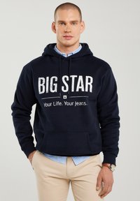 Vyras dėvintis tamsiai mėlynį džemperį su užrašu „BIG STAR Your Life. Your Jeans.“, po juo dėvi šviesiai mėlyną marškinių sluoksnį, smėlio spalvos kelnes ir riešo laikrodį.
