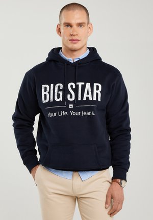 Mees, kes kannab tumesinist kapuutsiga sviitrit, millel on tekst "BIG STAR Your Life. Your Jeans.", seljas hele sinine särk, beežid püksid ja käekell.