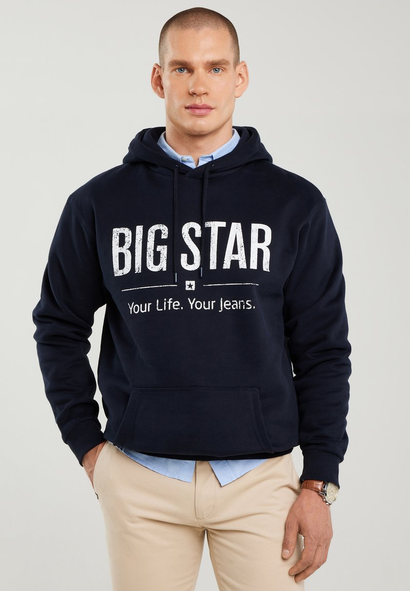 Vyras dėvintis tamsiai mėlynį džemperį su užrašu „BIG STAR Your Life. Your Jeans.“, po juo dėvi šviesiai mėlyną marškinių sluoksnį, smėlio spalvos kelnes ir riešo laikrodį.
