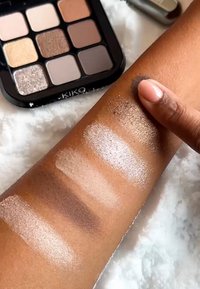 KIKO øyenskyggepalett med ni firkanter i myke nøytrale toner og metalliske farger; prøver påført en hudfarget overflate, som viser variert shimmer.