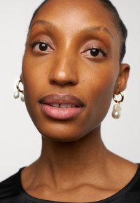 Svart topp med strukturerad tyg, gyllene hoops örhängen med vita pärlor. Slät hudton, naturlig sminkning, fokuserade ansiktsdrag.