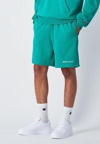 Türaquoise Shorts aus weichem Stoff mit elastischem Bund und Taschen; kombiniert mit weißen Sneakern und gerippten Socken mit einem Logo.
