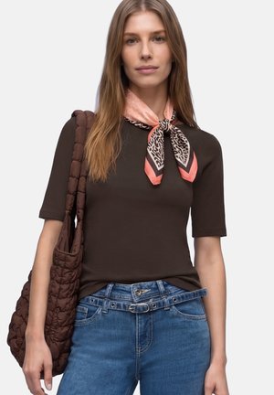 Frau mit schwarzem kurzärmligem Oberteil, rosa und animalprint Halstuch, blauen Jeans mit Gürtel, trägt eine große braune gesteppte Umhängetasche.