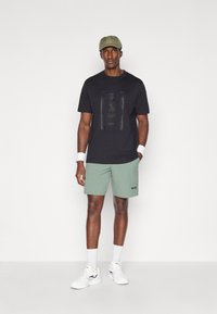 T-shirt noire avec un logo "BOSS" texturé, shorts verts avec impression "BOSS", chaussures de sport blanches et une casquette vert clair. Tissu lisse et coupe athlétique.