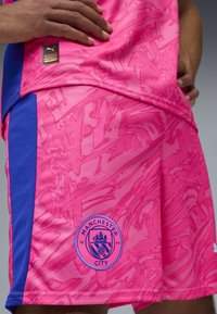 Różowy sportowy podkoszulek z abstrakcyjnym wzorem, z niebieskimi akcentami. Na dolnej części znajduje się logo Manchester City. Wykonany z oddychającego materiału.