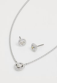 Set di gioielli in argento composto da una collana con catena e un ciondolo ovale, abbinata a orecchini a bottone, entrambi decorati con pietre preziose trasparenti.
