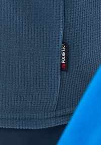 Dunkelblaues Fleece-Gewebe mit gerippter Textur, versehen mit einem Polartec-Label. Ein heller blauer Stoffteil ist teilweise im Hintergrund sichtbar.