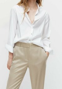Kobieta w luźnej białej koszuli zapinanej na guziki, włożonej w beżowe, wysokie spodnie, z jedną ręką w kieszeni, na neutralnym tle.