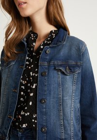 Giacca di jeans in un lavaggio medio con chiusura a bottoni, due tasche sul petto e cuciture a contrasto. Indossata sopra una maglietta nera a fiori.