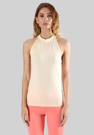 Femme aux cheveux ondulés portant un haut beige sans manches et un pantalon rose corail, debout devant un fond gris uni.