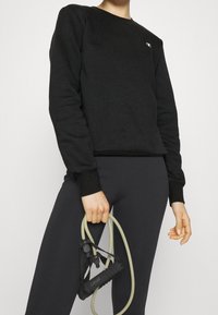 Felpa nera con maniche lunghe, scollo rotondo e logo. Abbinata a leggings neri. Il modello tiene in mano bande di resistenza per esercizi verdi.