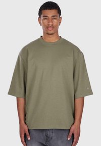 T-shirt oversize a maniche corte color verde oliva realizzato in cotone; presenta un colletto rotondo e un piccolo logo sulla manica sinistra.