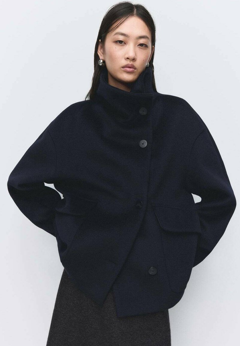 Chaqueta de lana azul marino con cuello alto, botones frontales y dos bolsillos laterales. Presenta un ajuste relajado y textura suave.