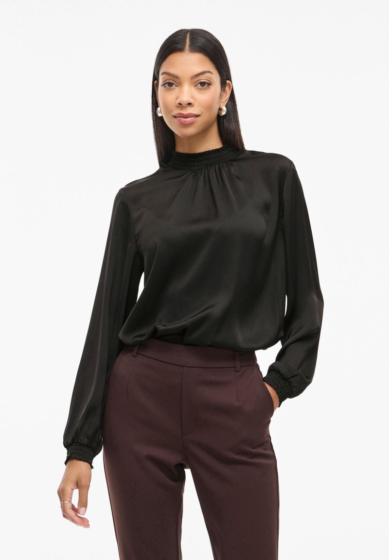 Schwarze Satinbluse mit langen Ärmeln, gerafftem Ausschnitt und elastischen Bündchen, kombiniert mit dunkelburgunderfarbenen, maßgeschneiderten Hosen. Glatte Textur insgesamt.