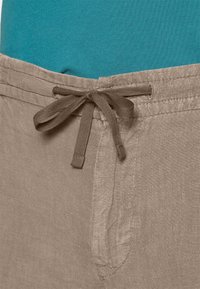 Ljusbruna linne-short med dragsko i midjan, med en strukturerad yta och sidofickor. Bakgrundsfärgen är teal.