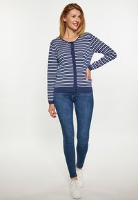 Cardigan a righe blu e bianche con collo rotondo e chiusura a bottoni, abbinato a jeans blu slim e sneakers bianche. Tessuto liscio.
