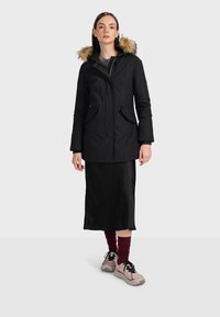 Čierna parka s kapucňou vystlanou umelým kožuchom, so zipsovým zapínaním, dvoma prednými vreckami a mierne priliehavým strihom. Nosená cez čierne šaty.